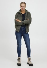 Groene fleece zip-up jack met hoge kraag, zwarte getailleerde shirt, blauwe skinny jeans en beige veterschoenen met zwarte zolen.
