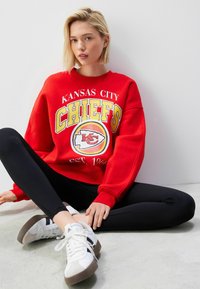 Piros kapucnis pulóver "Kansas City Chiefs" grafikával, sárga és fehér díszítéssel, fekete leggings-el és fehér, szürke akcentussal rendelkező sportcipőkkel párosítva.