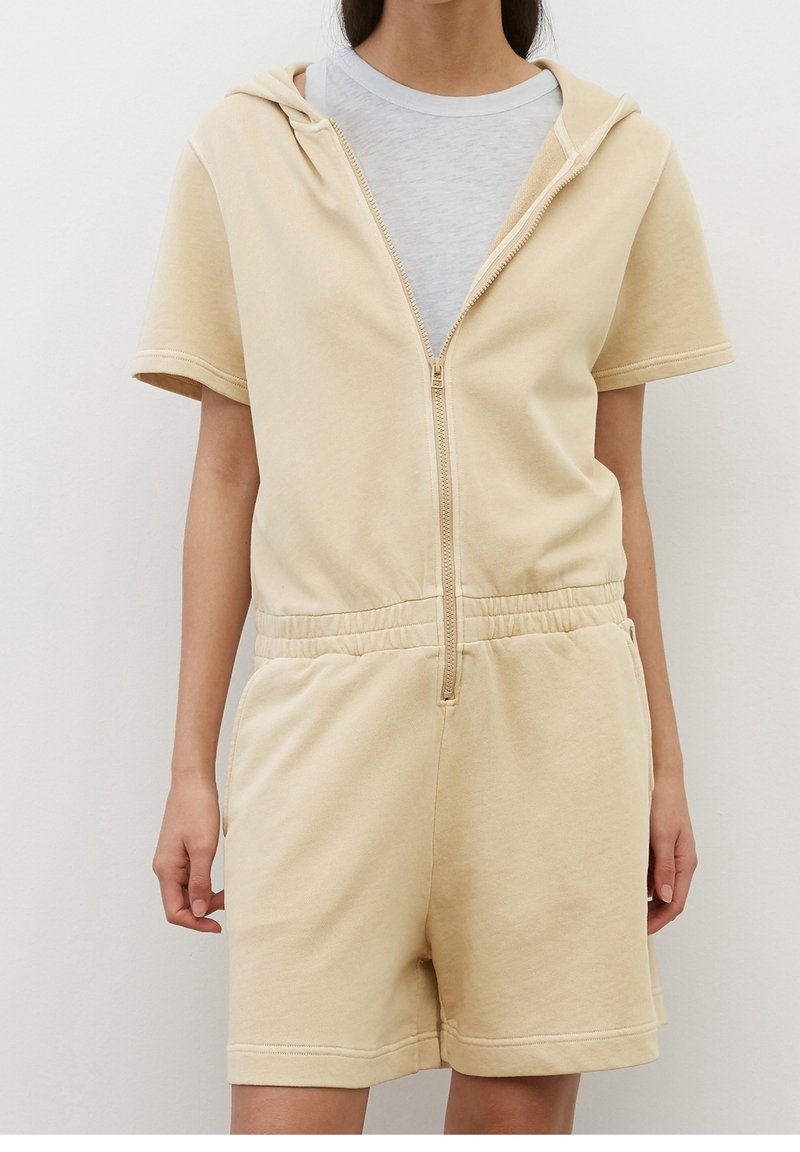 Femme portant une combinaison beige à manches courtes avec capuche, fermeture éclair sur le devant et taille élastique, par-dessus un t-shirt gris clair à col rond, debout sur un fond uni.