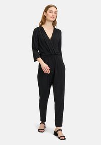Tuta nera con scollatura a V profonda, design drappeggiato, maniche a tre quarti e vestibilità morbida. Abbinata a sandali neri con tacco a blocco.