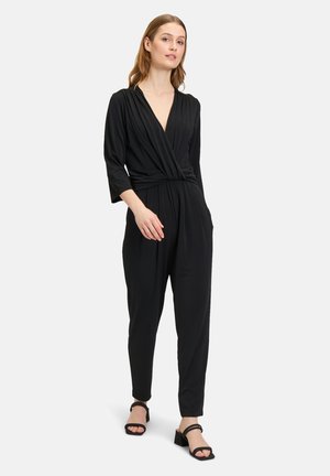 Tuta nera con scollatura a V profonda, design drappeggiato, maniche a tre quarti e vestibilità morbida. Abbinata a sandali neri con tacco a blocco.