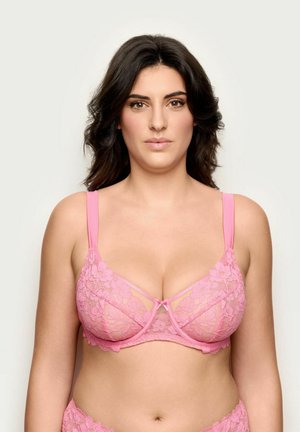 Kobieta o ciemnych falowanych włosach, ubrana w różowy koronkowy biustonosz, zwrócona twarzą do przodu na jednolitym jasnym tle.