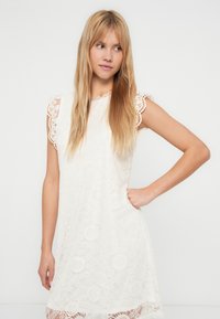 Balta mežģīņu kleita ar īsām, smalki izstrādātām piedurknēm un brīvu piegriezumu. Rakstaina mežģīņu kārta un viļņotas apakšmalas dizains.