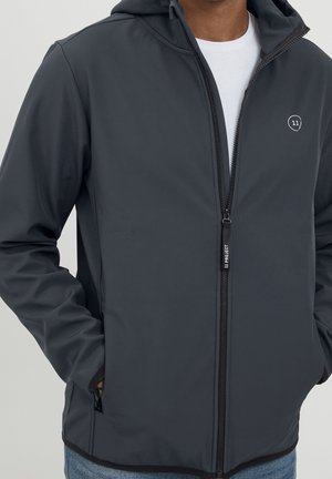 Mann trägt einen dunkelgrauen Kapuzenpullover mit Reißverschluss, Kapuze und kleinem weißem Logo auf der Brust, Hände in den Seitentaschen, darunter ein weißes Hemd und blaue Jeans.