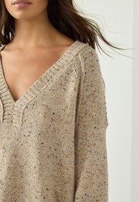 Beige strikket sweater med V-hals, struktureret mønster og subtile prikker af brunt og blåt. Drop-skulderdesign, blød finish.