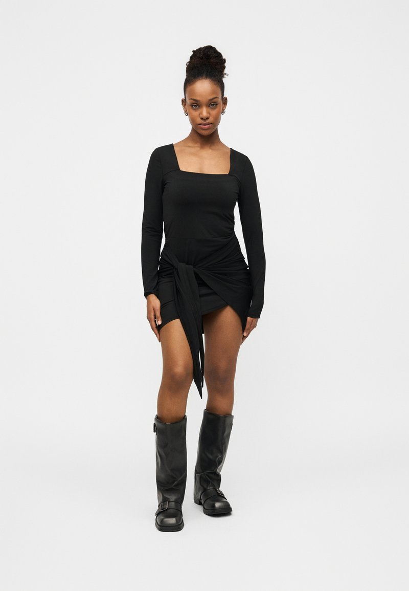 Μαύρο μίνι φόρεμα με μακριά μανίκια, τετράγωνο ντεκολτέ και λεπτομέρεια δέσιμου στο πλάι, συνδυασμένο με μαύρες δερμάτινες μπότες μέχρι το γόνατο. Ομαλή υφή.
