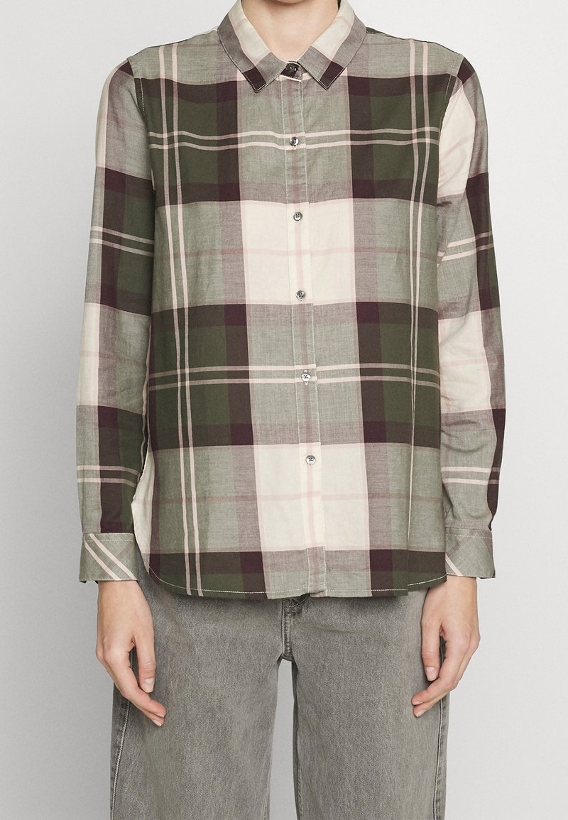 Personne portant une chemise à carreaux verte, blanche et marron à manches longues et un pantalon gris, debout devant un fond uni.