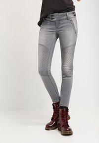 Vaqueros de denim gris con un corte ajustado, parches texturizados en las rodillas y cierre de doble botón; combinados con botas de combate rojas con cordones.
