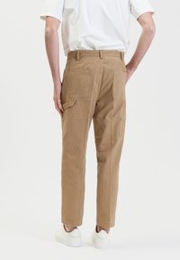 Bruine cargobroek van katoen, met een relaxte pasvorm, meerdere zakken en een soepele textuur. Draagt een eenvoudig wit t-shirt en schoenen.