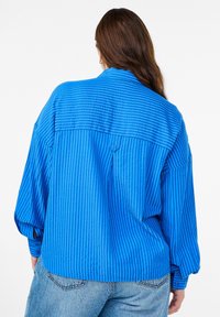 Žena s dlouhými hnědými vlasy, oblečená v volné modré pruhované košili a světlých modrých džínách, stojící zády k bílému pozadí.