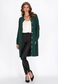 Sötétzöld kardigán, amelyet kábelmintázat és gyöngyökkel díszítettek, elöl gombos záródással. Világos felső és fekete leggings felett viselik.