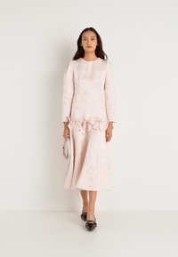 Μακρυμάνικο φόρεμα σε παστέλ ροζ απόχρωση με λουλουδάτο σχέδιο και ριγέ λεπτομέρεια στη μέση, συνδυασμένο με μαύρες μπαλαρίνες και μια μικρή ανοιχτό ροζ τσάντα.