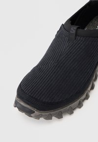 Melnas slip-on kurpes ar tekstūru auduma virspusi un izturīgu pelēku gumijas zoli, paredzētas āra vai ikdienas valkāšanai.