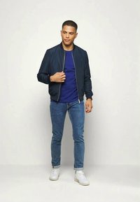 Casaco bomber azul-marinho sobre uma t-shirt azul, combinado com jeans azul-claro e ténis brancos. O tecido parece suave com um design clean.