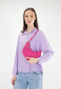Roze leren handtas met een gebogen vorm en ritssluiting, vastgehouden voor een model dat een lichtpaarse blouse en blauwe jeans draagt.
