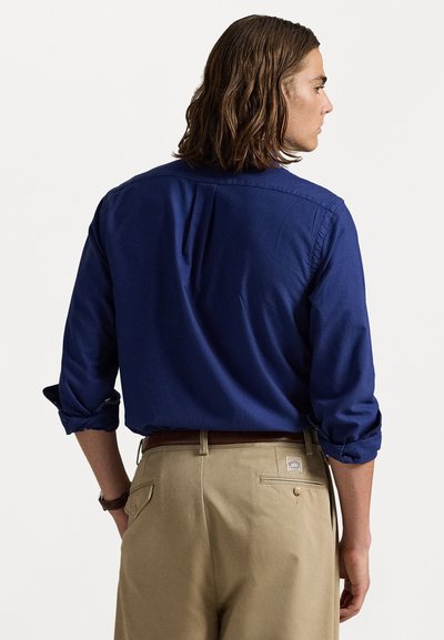 Tumši zila garām piedurknēm krekls ar pogājamiem manžetēm, kuram ir aizmugurējais izgriezums; apvienots ar gaiši khaki biksēm un brūnu jostu.