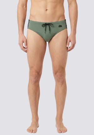 Uomo che indossa slip da bagno verde oliva con coulisse nera e loghi laterali, in piedi a piedi nudi su sfondo chiaro e semplice.