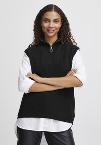 Schwarzer gestrickter ärmelloser Pullover mit hohem Kragen und Reißverschluss, versehen mit einem gerippten Muster. Getragen über einem weißen Hemd mit hochgekrempelten Ärmeln.