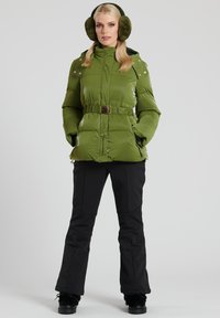Grön puffjacka med midjeskärp, glänsande textur och gyllen hardware. Kombineras med svarta snöbyxor och matchande fluffiga öronmuffar.