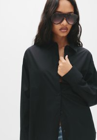 Camicia nera a maniche lunghe con colletto, in tessuto morbido, parzialmente aperta sul décolleté. Grandi occhiali da sole scuri coprono gli occhi.