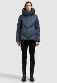 Sinine tepitud puffer-jope ševroontriibu mustriga, kõrge krae ja lukuga taskutega, mustad retuusid ja põlveni ulatuvad saapad halli taustal.