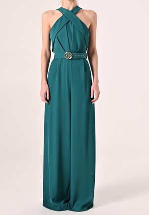 Femme portant une combinaison dos-nu bleu sarcelle avec des jambes larges et une ceinture décorative avec boucle ronde, debout devant un fond uni.