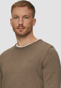 Brūns džemperis ar apaļu kakla izgriezumu un ribota aproce. Tam ir mīksta tekstūra un smalka logo detaļa uz krūtīm.