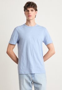 T-shirt azul claro de mangas curtas, feito de um tecido suave, com gola redonda e um discreto detalhe do logo no peito. Corte casual com uma textura lisa.