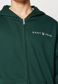 Sweat à capuche zippé vert en tissu doux, avec poche kangourou, capuche ajustable et logo brodé blanc "GANT 1949" sur la poitrine.