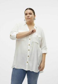 Hvid skjorte med knapper lavet af letvægtsstof, med to brystlommer, oprullede ærmer og trækknapper i træ. Båret med blå jeans.
