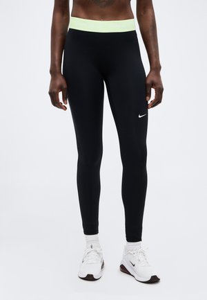 Person wearing black Nike leggings with a pale yellow waistband and white Nike sneakers, standing against a neutral background.