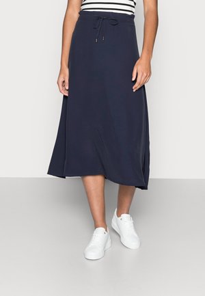 Femme portant une jupe midi bleu marine avec une taille à cordon et des baskets blanches sur un fond uni.