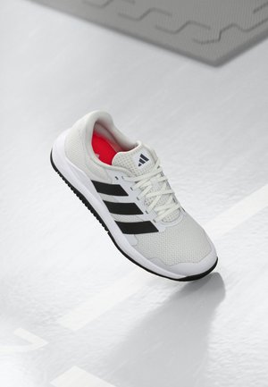 Biały but do biegania Adidas z czarnymi paskami i czerwonym wnętrzem, ustawiony pod kątem na błyszczącej szarej podłodze blisko linii boiska sportowego.