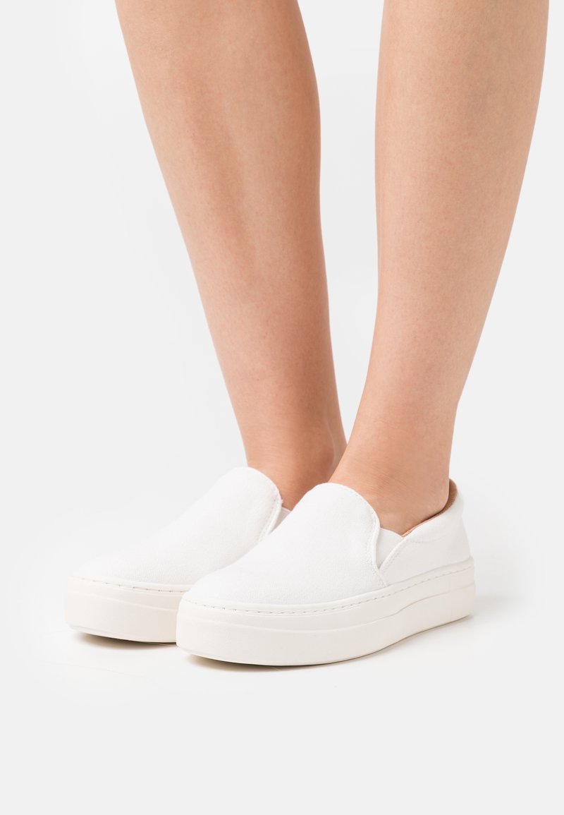 Pieds portant des baskets blanches unies à plateforme sans lacets avec une semelle épaisse sur un fond clair.