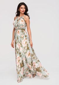 Femme portant une robe sans manches, à col halter, longue jusqu'au sol avec un motif floral pastel, debout et regardant légèrement sur le côté.