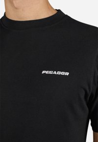 Sort bomulds T-shirt med tætsiddende pasform, med ribbet rund hals og ordet "PEGADOR" broderet i hvidt på forsiden.