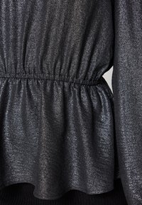 Haut noir scintillant avec une taille froncée et une forme fluide. Le tissu a un éclat métallique et une texture douce. Ourlet légèrement évasé.