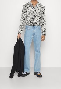 Zwarte bloemen blouse met lange mouwen, lichtblauwe flared jeans, houdt een zwarte trui vast en draagt zwarte schoenen met kleurrijke ontwerpen.
