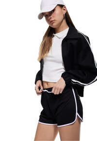 Chaqueta bomber negra con rayas blancas, camiseta polo blanca y pantalones cortos negros con ribete blanco. Los accesorios incluyen una gorra blanca y pendientes de aro.