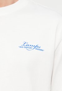 Λευκό πουκάμισο με μπλε κείμενο «Lamps 'Mid Century Modern'» τυπωμένο στο πάνω μέρος του στήθους.