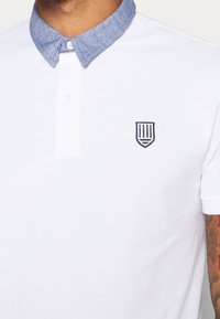 Polo blanc avec col en chambray bleu, deux boutons blancs, logo bouclier noir brodé sur la poitrine, et bras tatoué visible.
