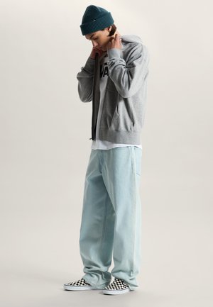 Młody mężczyzna w turkusowej czapce beanie, szarej bluzie z kapturem, białej koszulce Vans, jasnobłękitnych szerokich dżinsach oraz czarno-białych, w kratkę wsuwanych trampkach.