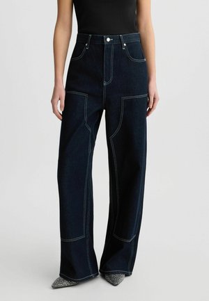 Jeans larges en denim foncé avec des coutures contrastantes, taille haute et deux poches avant. Offre un tissu subtilement texturé avec une coupe structurée.
