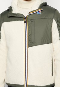 Grüne und cremefarbene Jacke mit Fleece- und Nylonstrukturen, ausgestattet mit einem Reißverschluss vorne, einer Kapuze und einem bunten Reißverschlussakzent.