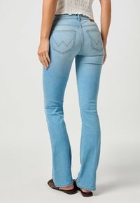 Jean flare bleu clair avec une texture en denim lisse, comportant deux poches arrière avec un design en zigzag et une taille mi-haute.