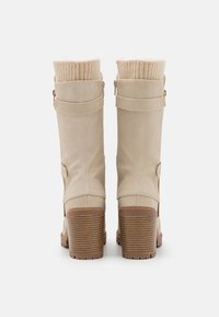 Botas de imitación de cuero beige hasta la rodilla con un puño de punto acanalado, detalles en tono dorado, cremalleras laterales y un tacón de madera apilada.