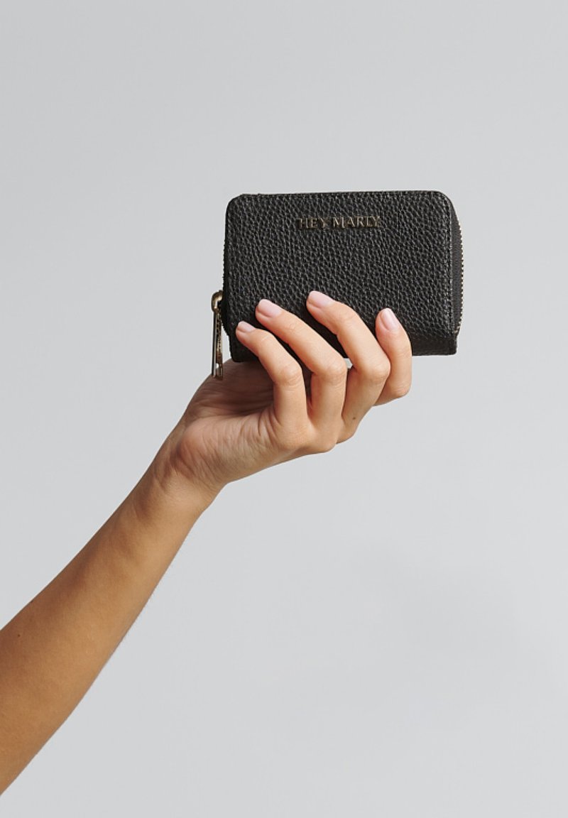 Zwarte leren portemonnee met textuur, vastgehouden in een hand, met een rits sluiting en een reliëf merknaam aan de voorkant.