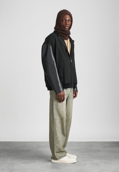 Giacca nera con zip e maniche in pelle, top beige, pantaloni verde palazzo e sneakers bianche; modello con un cappuccio di maglia marrone.