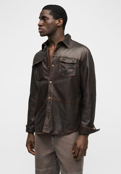 Νεαρός άνδρας που κοιτάζει προς τα αριστερά, φορώντας σκούρο καφέ δερμάτινο μπουφάν με μπροστινές τσέπες και γκρι παντελόνι, με απλό φόντο.