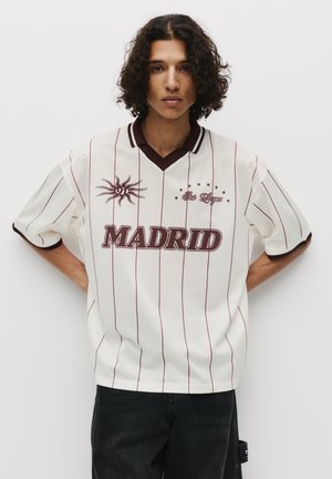 Maglia di mesh bianca con pinstripes bordeaux, scollatura a v e testo "MADRID". Presenta una grafica del sole e stelle sul davanti, maniche corte, vestibilità comoda.